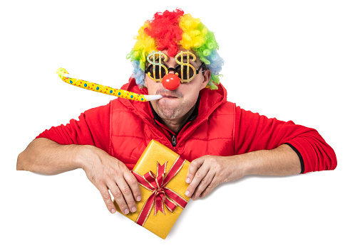 Funny clown in a multi colored wig with dollar shaped glasses and horn blower holding a gift box. Isolated on white. You can add extra white space with your message to the bottom.