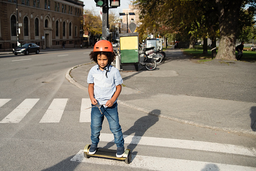 Mixed-race young boy on electric skateboard crossing street. He is wearing casual clothes and a protective helmet. Horizontal outdoors full length shot with copy space.