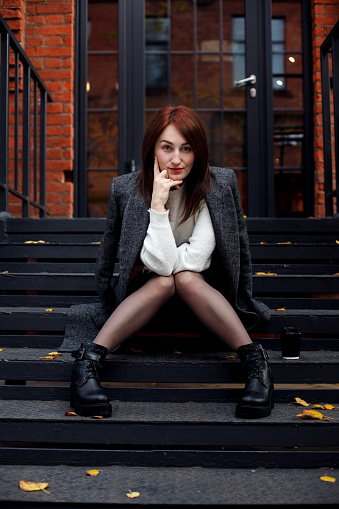 Attractive caucasian woman sits on a stair step and looks curiously at camera with elbows on her knees and chin on her hand. Fashion outdoor portrait at autumn street in New York style.