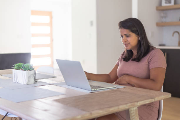 istock A thirty-something ethnic woman sits at her kitchen table in her home working. She is focused while looking at her laptop. 1281543145