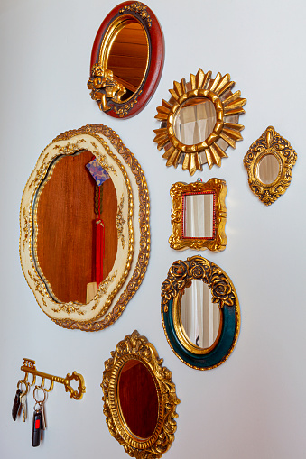 Bogota, Colombia - Gilded mirror frames and mirrors of different sizes and shapes neatly arranged and hung on a wall in the interior of a small apartment in the North part of the Andean capital city. Window curtains are reflected in some of the curtains while others reflect the front doors wooden panelling. Image shot in natural light; vertical format. No people.