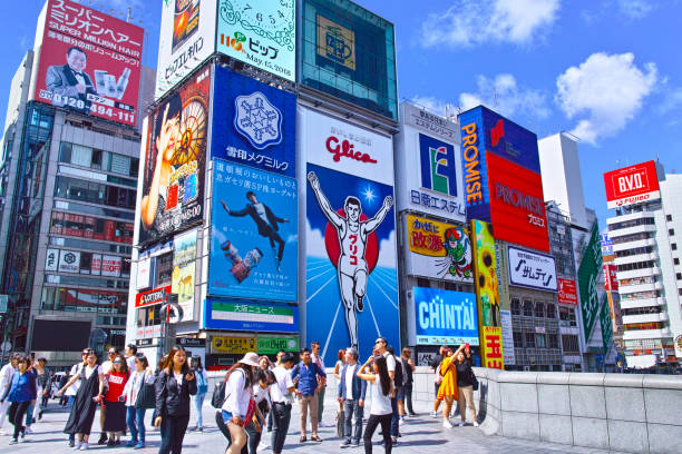 Oosaka, Japan-May 15, 2018: A view of Ebisubashi over the Dotonbori River in Chuo-ku, Osaka at noon. The runner signboard of the confectionery company seen from this bridge is famous, and the bridge is crowded with tourists. In the photo, tourists take pictures against the background of this sign, and a group of travelers are chatting.