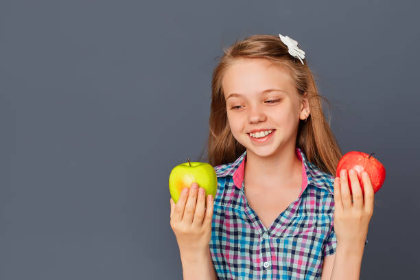 una niña linda elige entre una manzana verde y roja sobre un fondo gris. el concepto de elegir o tomar una decisión difícil - decisions teenage girls horizontal studio shot fotografías e imágenes de stock