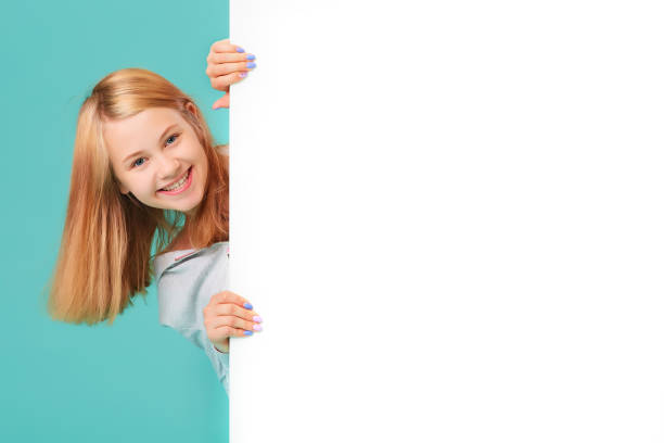 a laughing girl standing against a turquoise background holds an empty white Board in her hand and looks out from behind it a laughing girl standing against a turquoise background holds an empty white Board in her hand and looks out from behind it. peeking photos stock pictures, royalty-free photos & images