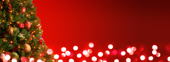 A Christmas tree full of ornaments and lights in front of a red background and blurred lights.  The negative space provides ample room for copy and text.