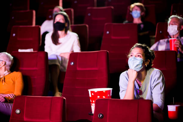Young woman and the other spectators wearing protective face masks at the cinema Audience at the cinema wearing protective face masks and sitting on a distance while watching the movie. movie theater photos stock pictures, royalty-free photos & images