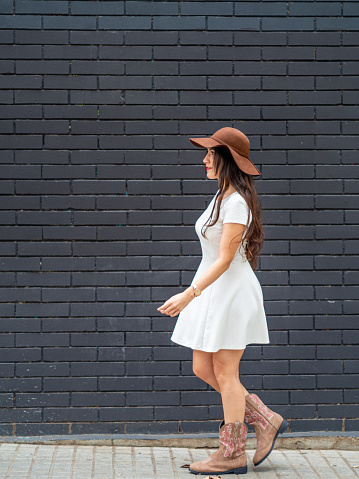 Joven con un vestido blanco, sombrero y botas de vaquero caminando frente a una pared de ladrillo negro. photo