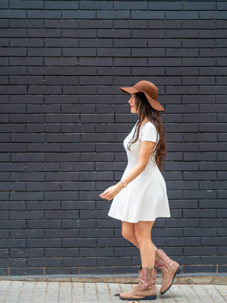 young woman wearing a white dress, hat and cowboy boots walking in front of a black brick wall. - bangs fashion model women elegance imagens e fotografias de stock