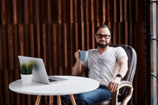 istock Young successful bearded businessman freelancer in office coworking. Happy man programmer developer in a white t shirt smiling and drinking coffee at the Desk with a laptop 1281594114