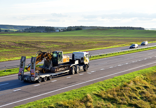 Trailer truck with long platform transport the Excavator on highway. Earth mover backhoe on heavy duty flatbed vehicle for transported. Long Haul and Shipping Services. Loading on Low Bed Trailer