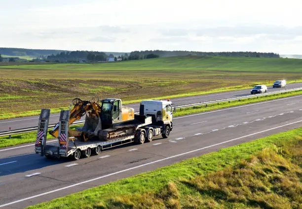 Photo of Trailer truck with long platform transport the Excavator on highway. Earth mover backhoe on heavy duty flatbed vehicle for transported.