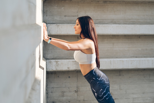 Sporty young woman doing standing wall push up triceps strength exercise during urban outdoor fitness workout.