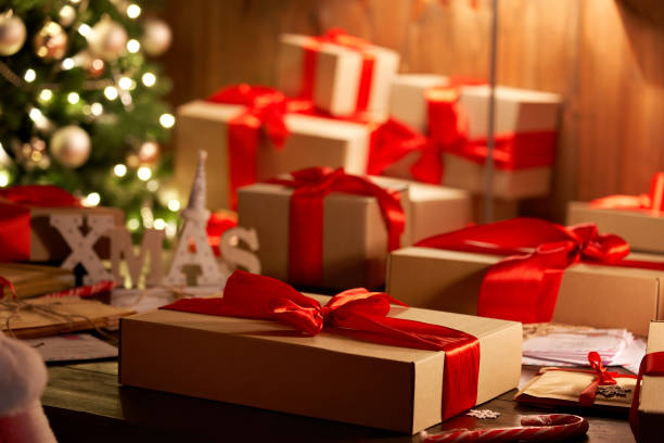 istock Merry Christmas gift on decorated festive table in cozy Santa workshop interior with xmas tree. Present wrapped with red ribbon, Happy New Year box in late night lamp light on holiday eve background. 1281596604