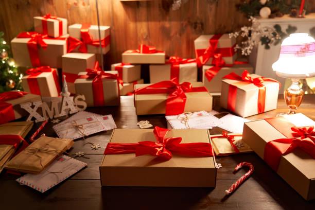 istock Wrapped Merry Christmas gift on decorated festive table in cozy Santa workshop interior with xmas tree. Present with red ribbon, Happy New Year box in late night lamp light on holiday eve background. 1281596607