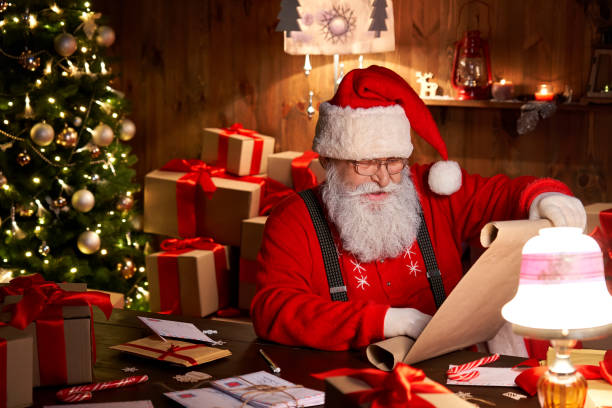 feliz viejo barbudo santa claus usando sombrero sosteniendo pergamino rollo leyendo la lista de deseos de navidad sentado en la acogedora mesa del taller casero tarde con regalos, árbol y velas preparándose para las vacaciones en la víspera de navidad. - santa hat christmas hat headwear fotografías e imágenes de stock