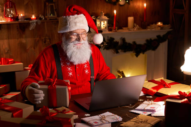glückliche alte weihnachtsmann trägt hut halten geschenk-box mit laptop-computer sitzt am werkstatt-heimtisch spät am frohen weihnachtsabend. e-commerce-website xmas zeit urlaub online-shopping e-commerce-verkauf - nikolaus stock-fotos und bilder