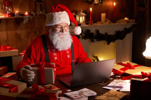 Happy old Santa Claus wearing hat holding gift box using laptop computer sitting at workshop home table late on Merry Christmas eve. Ecommerce website xmas time holiday online shopping e commerce sale