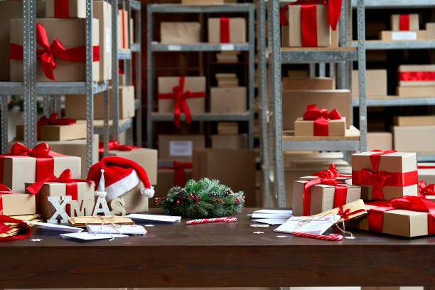 istock Decorated Merry Christmas table with gifts boxes in warehouse interior background. Many presents wrapped with red ribbons and letters on desk in storage. Xmas postal shipping delivery concept. 1281596614
