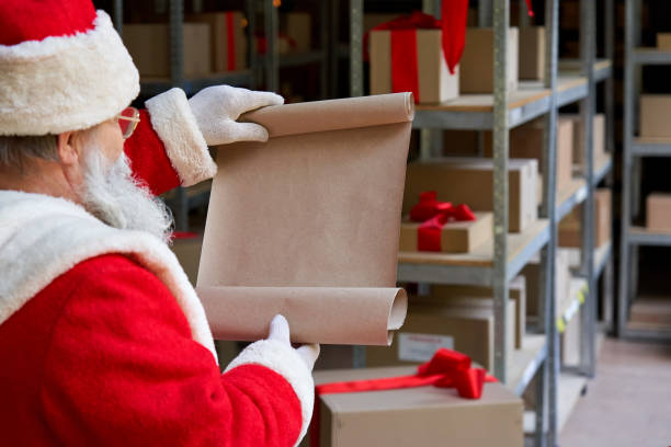 Over shoulder view of old Santa Claus wearing costume reading blank empty wish list standing in workshop gifts warehouse. Merry Christmas wishlist presents parcels post shipping delivery concept. sobre la vista del hombro del viejo santa claus usando el traje leyendo la lista de deseos vacía en blanco de pie en el almacén de regalos del taller. feliz lista de deseos de navidad presenta paquetes después del concepto de entrega de envío.