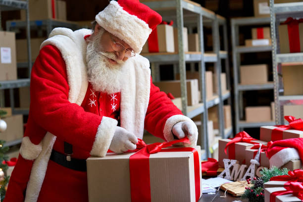 istock Happy old bearded Santa Claus, Saint Nicholas packing present box wrapping gift with red ribbon preparing post shipping delivery parcel on table in workshop. Merry Christmas shipping delivery concept. 1281596619
