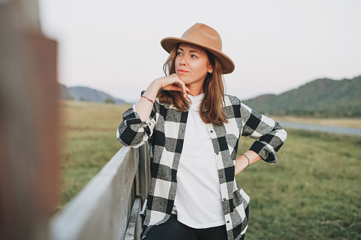Young woman in felt hat and plaid shirt near old wooden fence on mountains background