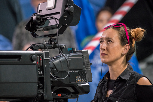Camerawoman looking at camera while shooting basketball match in stadium.