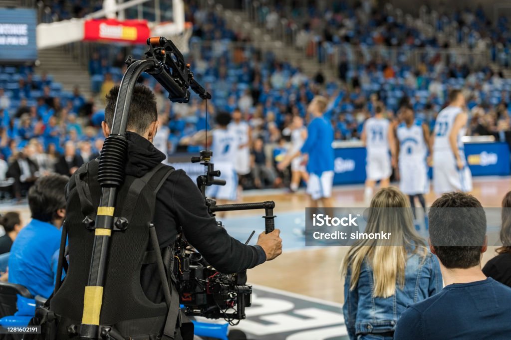Cameraman operating camera in court Rear view of cameraman shooting basketball match in stadium. Sport Stock Photo