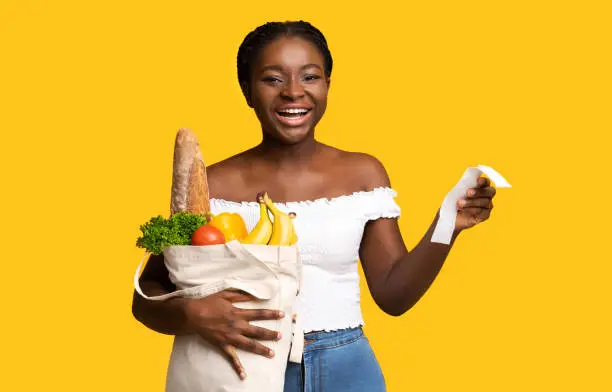 Photo of Cheerful black woman holding supermarket bill and bag with groceries, yellow background