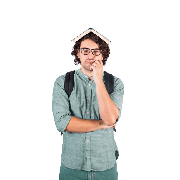 istock Puzzled and perplexed student guy keeps hand under cheek, holding a book over head, looks exhausted of constant learning. Young man brunette with long curly hairstyle wears glasses isolated on white. 1281610776