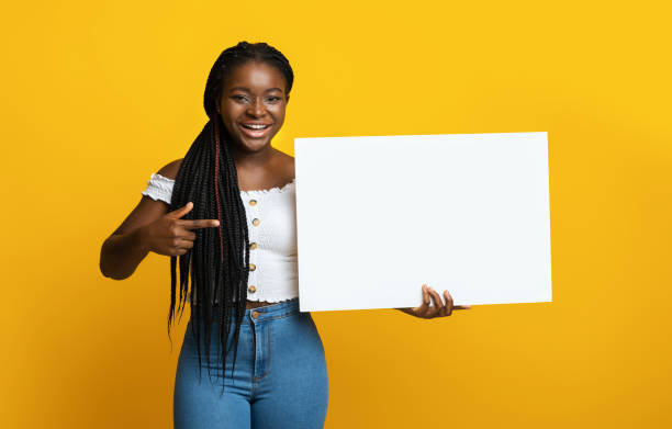 allegra signora nera che tiene e punta a un cartello bianco bianco, sfondo giallo - cardboard sign foto e immagini stock