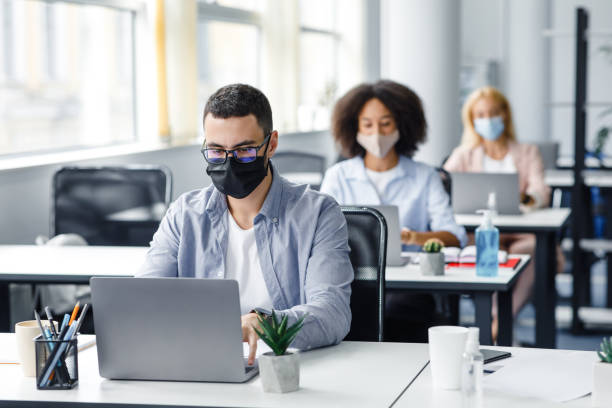 istock Teamwork in corporate company and returning to work after quarantine covid-19. Focused millennial man in glasses and protective mask works at laptop at workplace with antiseptic 1281613756