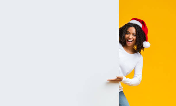 Cheerful Afro Woman In Santa Hat Pointing At White Advertisement Board