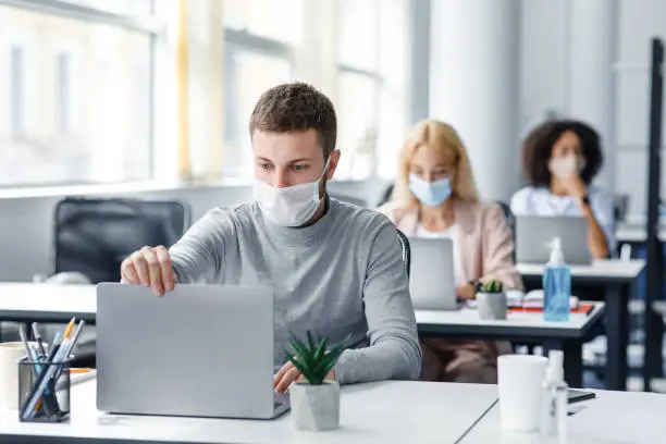 Photo of Coronavirus quarantine and office work with colleagues keeping social distancing. Portrait of young man in protective mask make video call with client