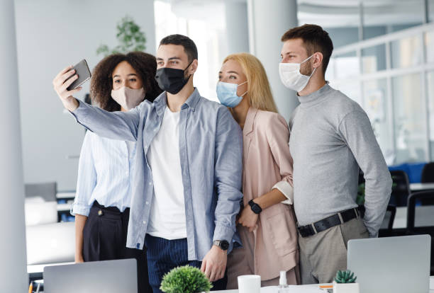istock Corporate selfies, team building and new normal during COVID-19 outbreak. Multiracial workers in protective masks makes photo with smartphone in interior of modern office 1281616375