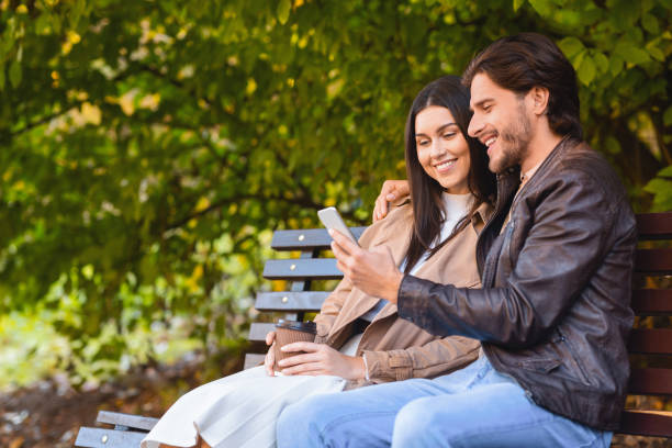 glückliches paar sitzt auf bech im park, mit handy - bech stock-fotos und bilder