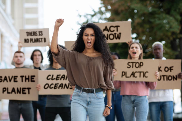 Young woman raising fist up, leading group of demonstrators Save the nature. Mixed race young woman raising fist up and yelling, leading group of demonstrators on the street. Multiracial group of young people with placards protesting for healthy environment greenpeace activists stock pictures, royalty-free photos & images