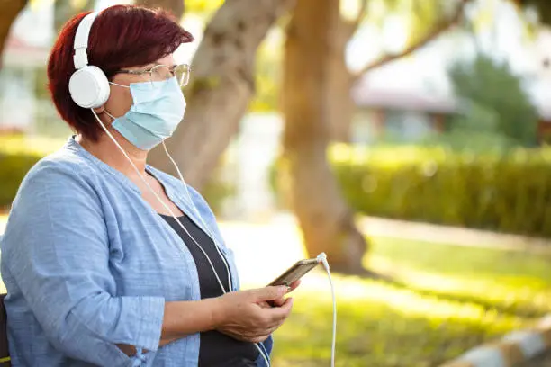 Photo of Mature woman with phone and headphones. mature woman in medical mask new normal socialization concept.