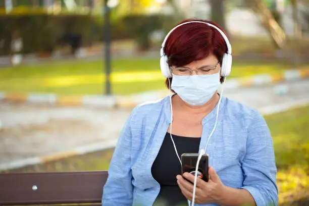 Photo of Mature woman with phone and headphones. mature woman in medical mask new normal socialization concept.