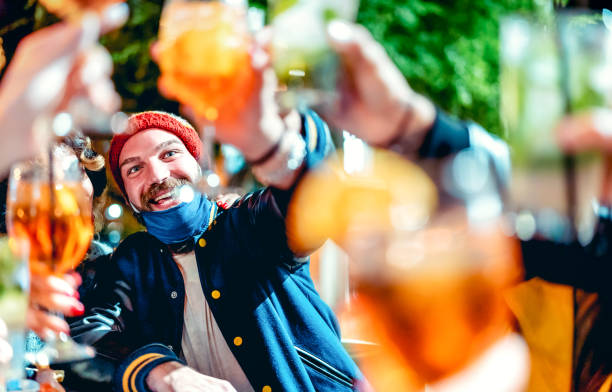 Happy guy toasting fancy drinks at night bar with open mask - New normal lifestyle concept with  friends having fun together on winter clothes - Focus on face with defocused background and foreground Happy guy toasting fancy drinks at night bar with open mask - New normal lifestyle concept with  friends having fun together on winter clothes - Focus on face with defocused background and foreground apres ski stock pictures, royalty-free photos & images