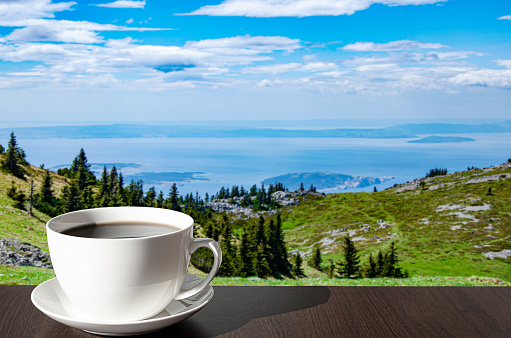 Cup of coffee on the table against mountains background. View of blue sea and green valley in the mountains.