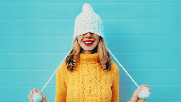ritratto invernale di donna allegra tira un cappello sugli occhi indossando maglione a maglia gialla e cappello bianco con pom pom su sfondo blu - golf women surprise playing foto e immagini stock