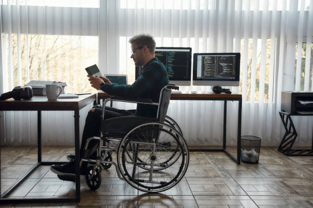 istock Such an interesting book. Side view of young male web developer in a wheelchair holding a book while sitting at his workplace with multiple computer screens on the background 1281642545