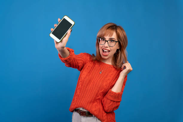 istock Image of excited young woman making winner gesture and using cellphone isolated over blue background. 1281644831