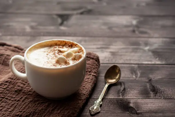 White cup of cappuccino with marshmallows and spoon on a wooden table. Selective focus, copy space