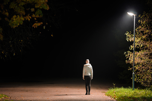 Young adult alone woman in white jacket standing on road under lamp light in autumn night. Back view. Road to nowhere concept. Empty place for text, quote or sayings.