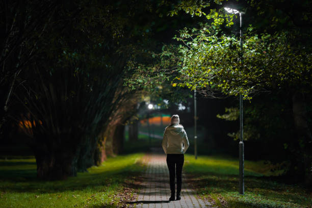 One young alone woman in white jacket walking on sidewalk through alley of trees under lamp light in autumn night. Spending time alone in nature. Peaceful atmosphere. Back view. One young alone woman in white jacket walking on sidewalk through alley of trees under lamp light in autumn night. Spending time alone in nature. Peaceful atmosphere. Back view. dark street stock pictures, royalty-free photos & images