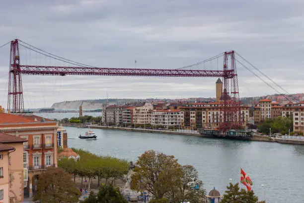Portugalete, Spain - 10/17/2019: Biscay bridge flying with gondola and boat on river Nervion, toned. Portugalete landmark. Famous bridge called Puente de Vizcaya near Bilbao. Riverbank of Portugalete in autumn.
