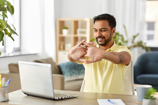 technology, remote job and lifestyle concept - happy smiling indian man with laptop computer stretching arms at home office