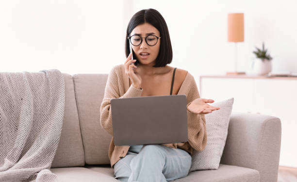 istock Frustrated asian woman talking on mobile phone sitting on sofa 1281654694