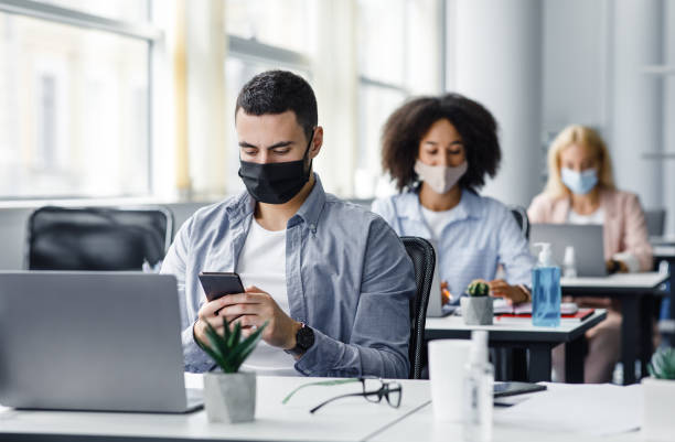 istock Modern gadgets for working with clients remotely from office after quarantine. Arabic young guy in protective mask typing on smartphone at table with laptop 1281654745